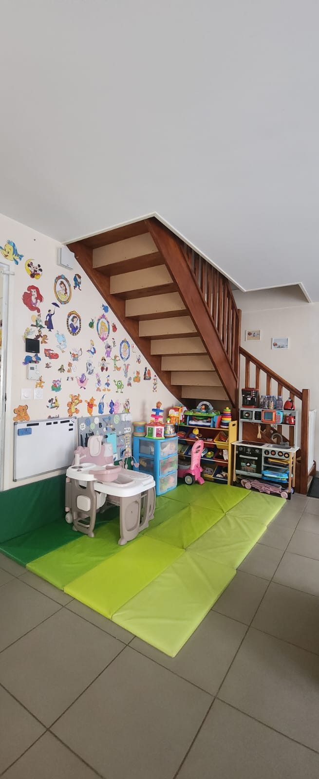 Coin jeux sous l'escalier : tapis de motricité verts, étagères de jouets d'éveil, dînette, tableau d'activités